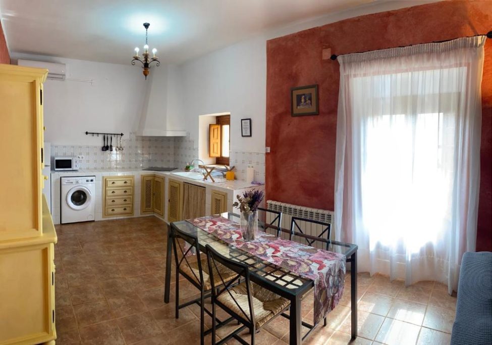 Cocina luminosa con muebles amarillos, mesa para cuatro y ventana con cortinas blancas.