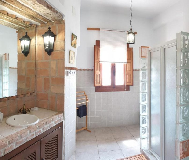 Baño luminoso con decoración rústica, lavabo, ducha y ventana con contraventanas abiertas.