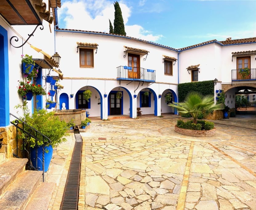 Patio con arquitectura blanca y azul, plantas y suelo de piedra. Cielo despejado.